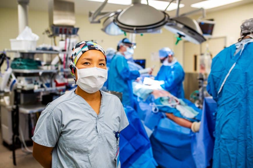 A nurse stands in the foreground of an operating room with other healthcare staff.